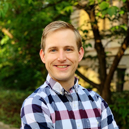 A professional photo of Andy Tonsing shows him standing in front of a blurred natural background with trees and sunlight. He is wearing a plaid shirt and smiling, embodying the dynamic presence of an Impact Summit leader. He has short blonde hair that is styled to the side.