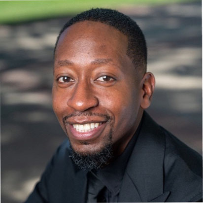 A professional photo of Daniel Clarke smiling confidently at the camera in an outdoor setting, embodying the dynamic presence of an Impact Summit leader. He wears a black business suit and has close-cropped hair. He has a polished beard and mustache.