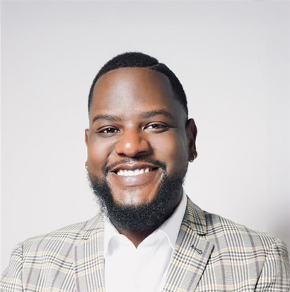 A professional photo of Darvelle Hutchins. He is sitting in front of a light grey backdrop and smiling confidently, embodying the dynamic presence of an Impact Summit leader. He wears a plaid blazer with a white button-up underneath. He has a short beard and close-cropped hair.