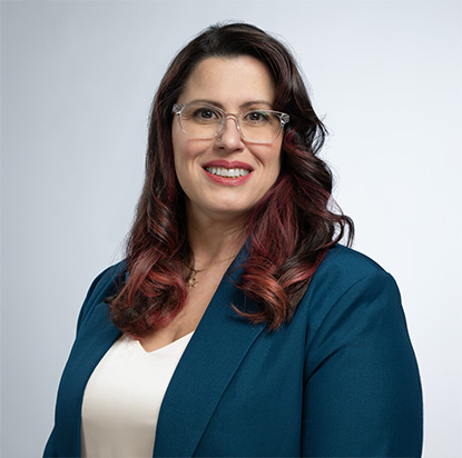 A professional photo of Donna Bungard. She is confidently standing in front of a light grey backdrop and smiling, embodying the dynamic presence of an Impact Summit leader. She wears a blue blazer with a white blouse underneath. She has long, curled brown hair with red highlights.