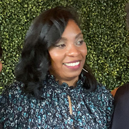 A photo of Jennifer Milliner-Bracey at an event. She is standing in front of a natural green backdrop, smiling, and looking off to the side. She wears a floral-printed blouse with variations of blue with a scallop trim at the neckline. She also has shoulder-length, dark brown, curled hair and radiates the dynamic presence of an Impact Summit leader.