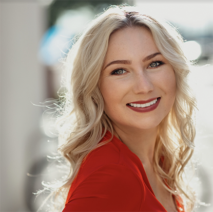 A professional photo of Keely Cat-Wells outdoors in natural sunlight and smiling confidently at the camera. She wears a red blouse, has long, wavy blonde hair, and embodies an Impact Summit leader's poised, dynamic presence.