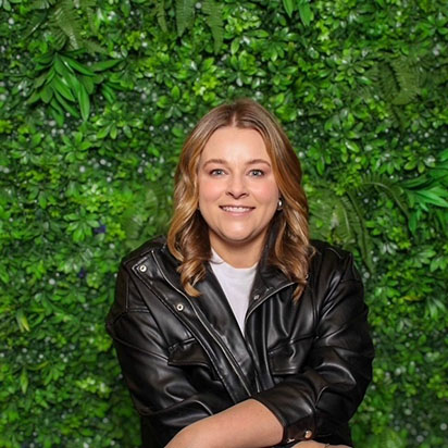 A cheerful photo of Kristen Lee captures her standing in front of a vibrant, leafy green background. She smiles confidently, embodying the dynamic presence of an Impact Summit leader. Kristen has short, wavy dark blonde hair and wears a black moto jacket over a white shirt.