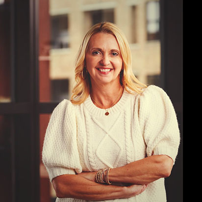 A professional photo of Lisa Schumacher features her standing confidently with folded arms. She smiles warmly, with a building blurred in the background. She has long blonde hair and wears a white sweater. She embodies the professional and confident demeanor of an Impact Summit leader.