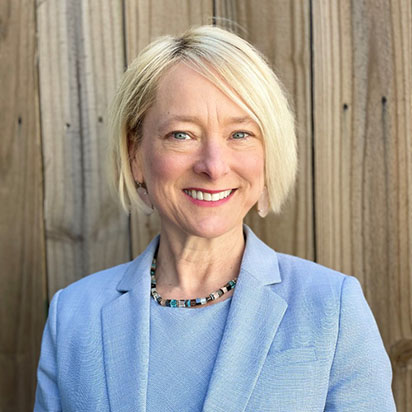 A professional photo of Missy Hopson smiling warmly in front of a wooden backdrop. She wears a light blue blazer, a beaded necklace, and has a short blonde bob. She reflects the confident, welcoming presence of an Impact Summit leader.