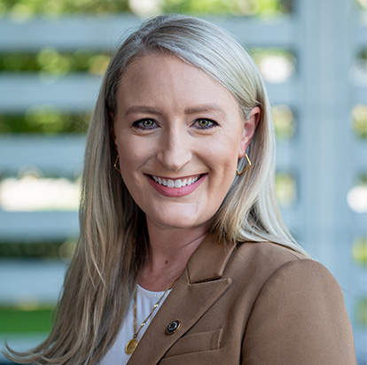 A professional photo of Paige Carter smiling confidently at the camera in an outdoor setting. She wears a tan blazer over a white top, with long, straight blonde hair and gold accessories. She embodies the dynamic presence of an Impact Summit leader.