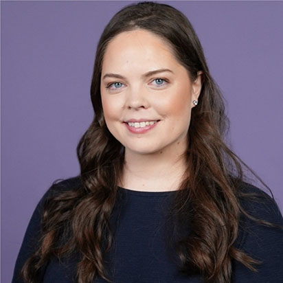 A professional photo of Rebecca Hinds. She sits in front of a solid purple background, smiling and looking at the camera confidently. She has long brown, curled hair and wears a dark top. Her expression conveys warmth and embodies the dynamic presence of an Impact Summit leader.