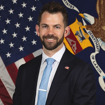 A professional photograph of Taylor Stockton shows him wearing a suit with a light blue tie and an American flag pin. He smiles confidently, embodying the dynamic presence of an Impact Summit leader. He has short brown hair and a well-kept beard and mustache. In the background, the U.S. flag and the Department of Labor flag are prominently displayed.