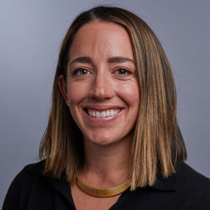 A professional photograph of Zoe Weintraub Barrett features her seated against a neutral grey backdrop. She is smiling confidently at the camera. Zoe has shoulder-length brown hair with golden highlights and is dressed in a black top, complemented by a gold necklace. Her expression and attire convey a professional demeanor, embodying the qualities of an Impact Summit leader.